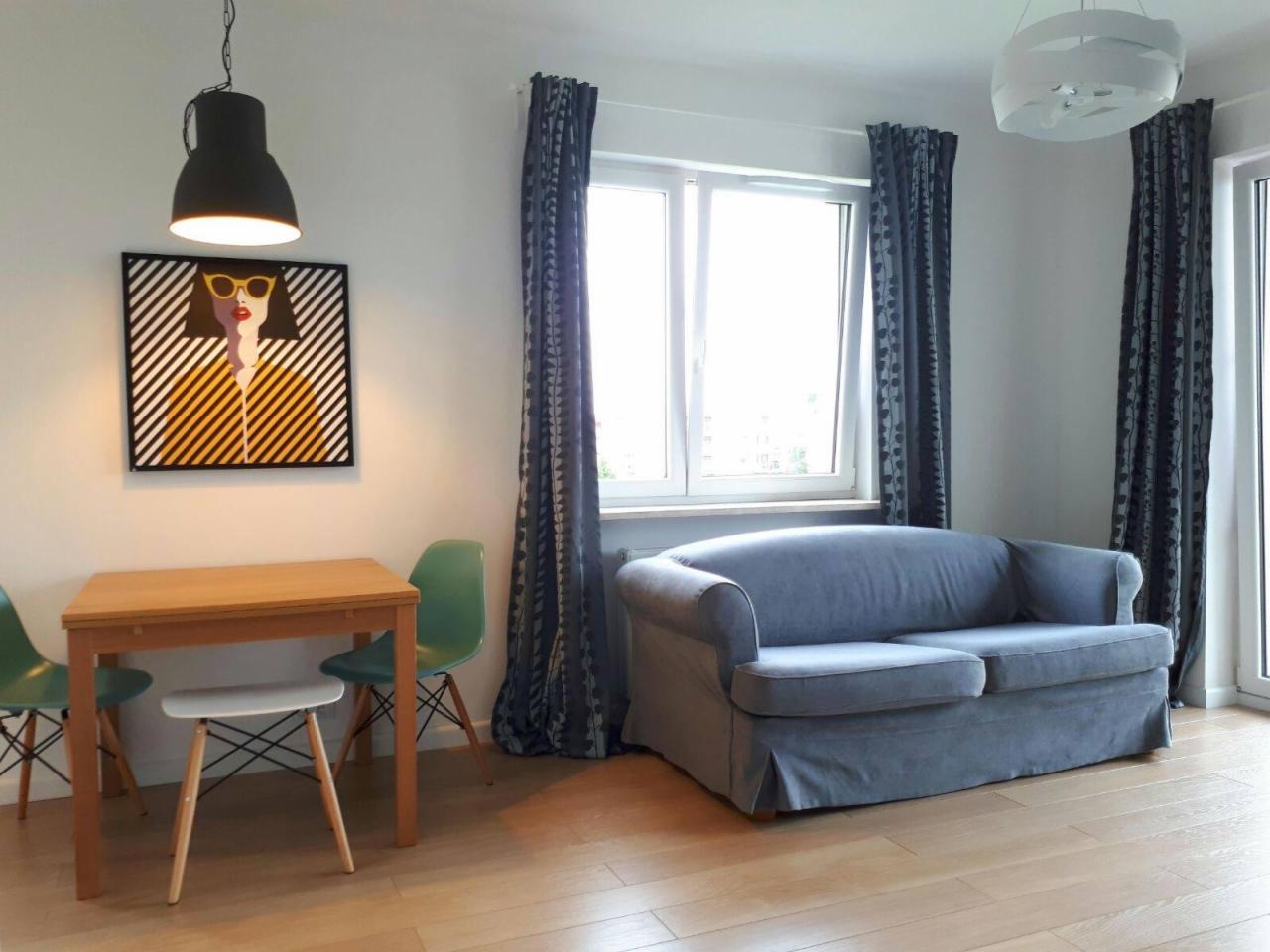 Jasne wnętrze pokoju dziennego z drewnianą podłogą, szarą sofą, zasłonami w kwiatowy wzór, drewnianym stołem z krzesłami i nowoczesnym obrazem na ścianie