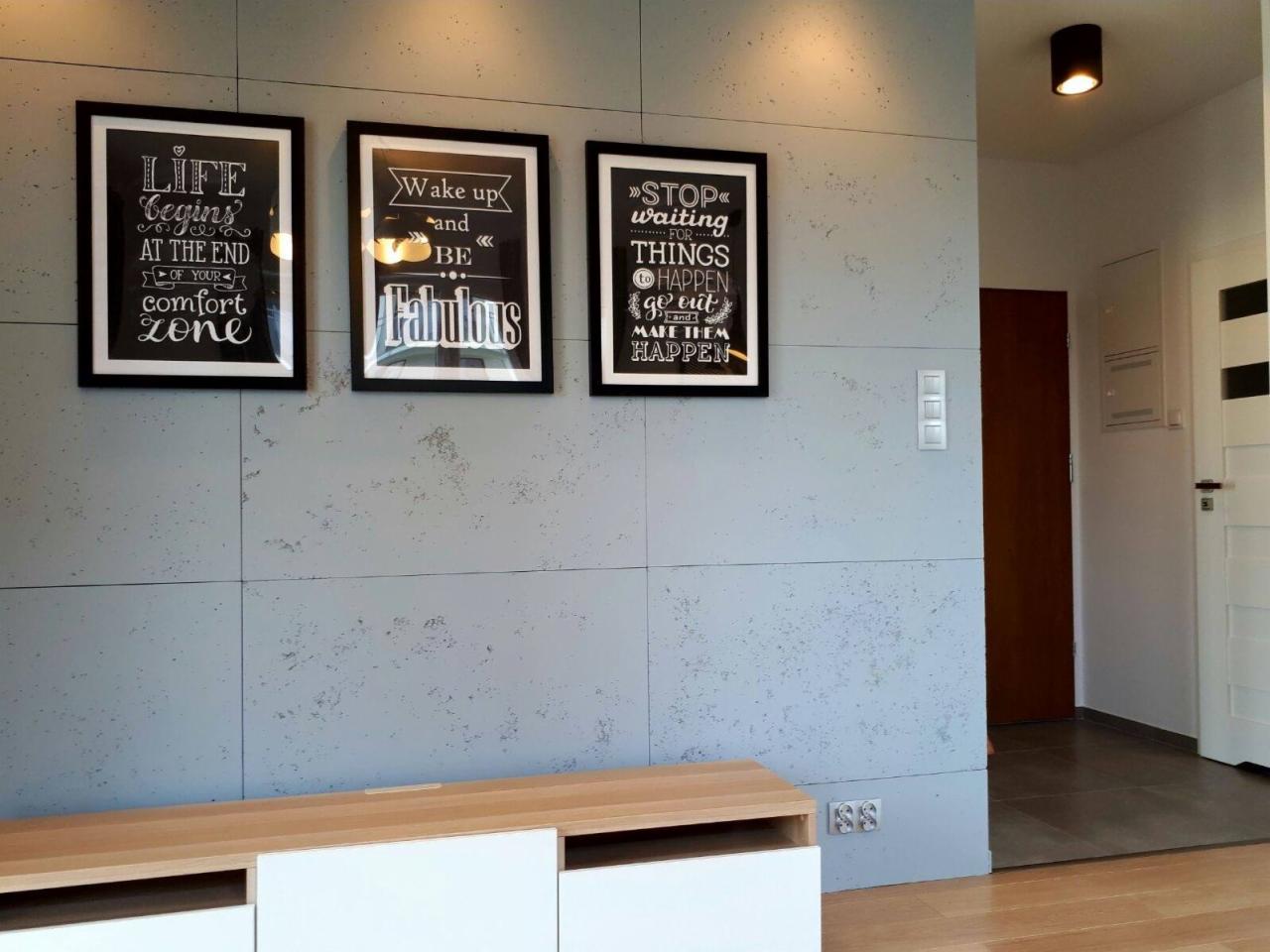 Szara ściana z betonową fakturą, ozdobiona trzema czarnymi ramkami z motywacyjnymi napisami, nad niską szafką RTV z białymi frontami, w tle widoczne drzwi wejściowe i oświetlenie sufitowe.