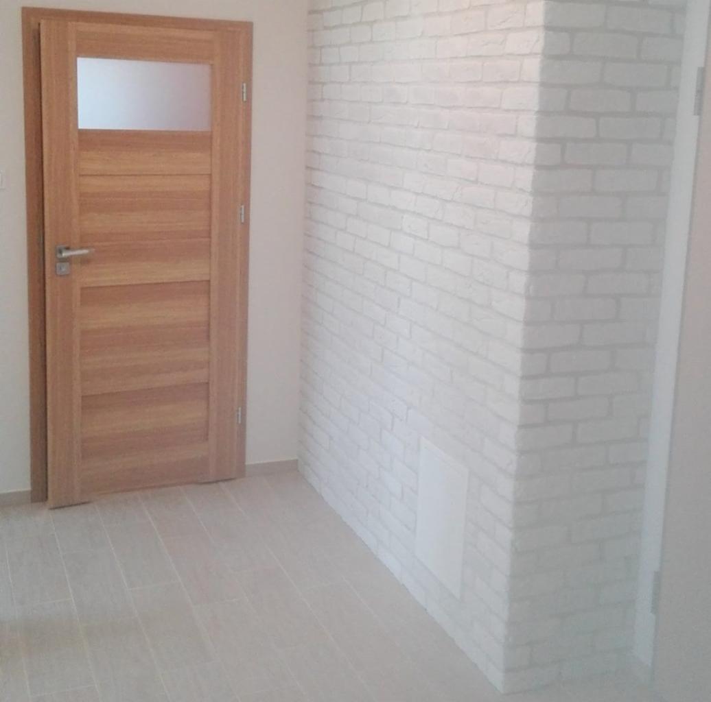 Jasne wnętrze po remoncie: drewniane drzwi z szybą, ściana z białej cegły i podłoga z płytek imitujących drewno.