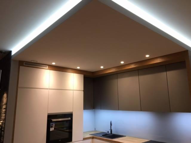 Nowoczesna kuchnia z białymi i grafitowymi szafkami, drewnianym blatem, oświetleniem LED podszafkowym i sufitowym, oraz wbudowanym piekarnikiem.