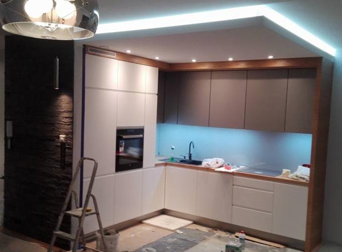 Nowoczesna kuchnia z białymi i szarymi szafkami, oświetleniem LED i czarną armaturą, widoczna drabina malarska i ślady po remoncie na podłodze.