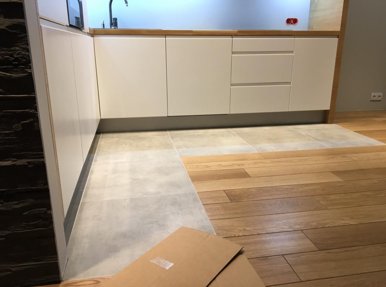 Nowoczesna kuchnia z białymi szafkami i blatem z jasnego drewna; podłoga łączy szare płytki i drewniane panele; fragment kartonu na pierwszym planie.