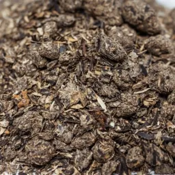 Stos szarej, kruszącej się zaprawy z widocznymi kawałkami drewna na białym tle, ukazujący strukturę mieszanki.