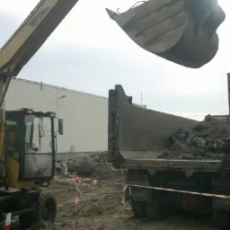 Żółta koparka CAT ładuje gruz do wywrotki SCANIA na placu budowy. Widoczne taśmy zabezpieczające teren. Hala w tle.