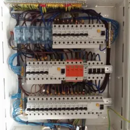 Otwarta skrzynka bezpiecznikowa z licznymi przewodami w różnych kolorach, widoczne zabezpieczenia i listwy zaciskowe, instalacja w trakcie podłączania.