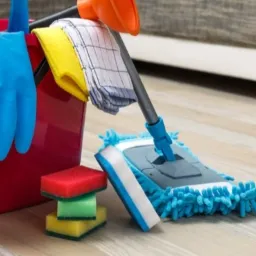 Czerwone wiadro z niebieskimi gumowymi rękawicami, ściereczkami i gąbkami do czyszczenia, obok mop z frędzlami na drewnianej podłodze w pokoju.