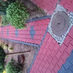 Ułożona z czerwono-szarych kostek ścieżka ogrodowa z widoczną studzienką kanalizacyjną otoczoną kostką brukową, otoczona zielenią.