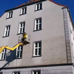 Pracownik w żółtym kombinezonie w koszu podnośnika na tle szarej, wielopiętrowej kamienicy z widocznymi zabrudzeniami na elewacji, wskazującymi na trwający proces czyszczenia.