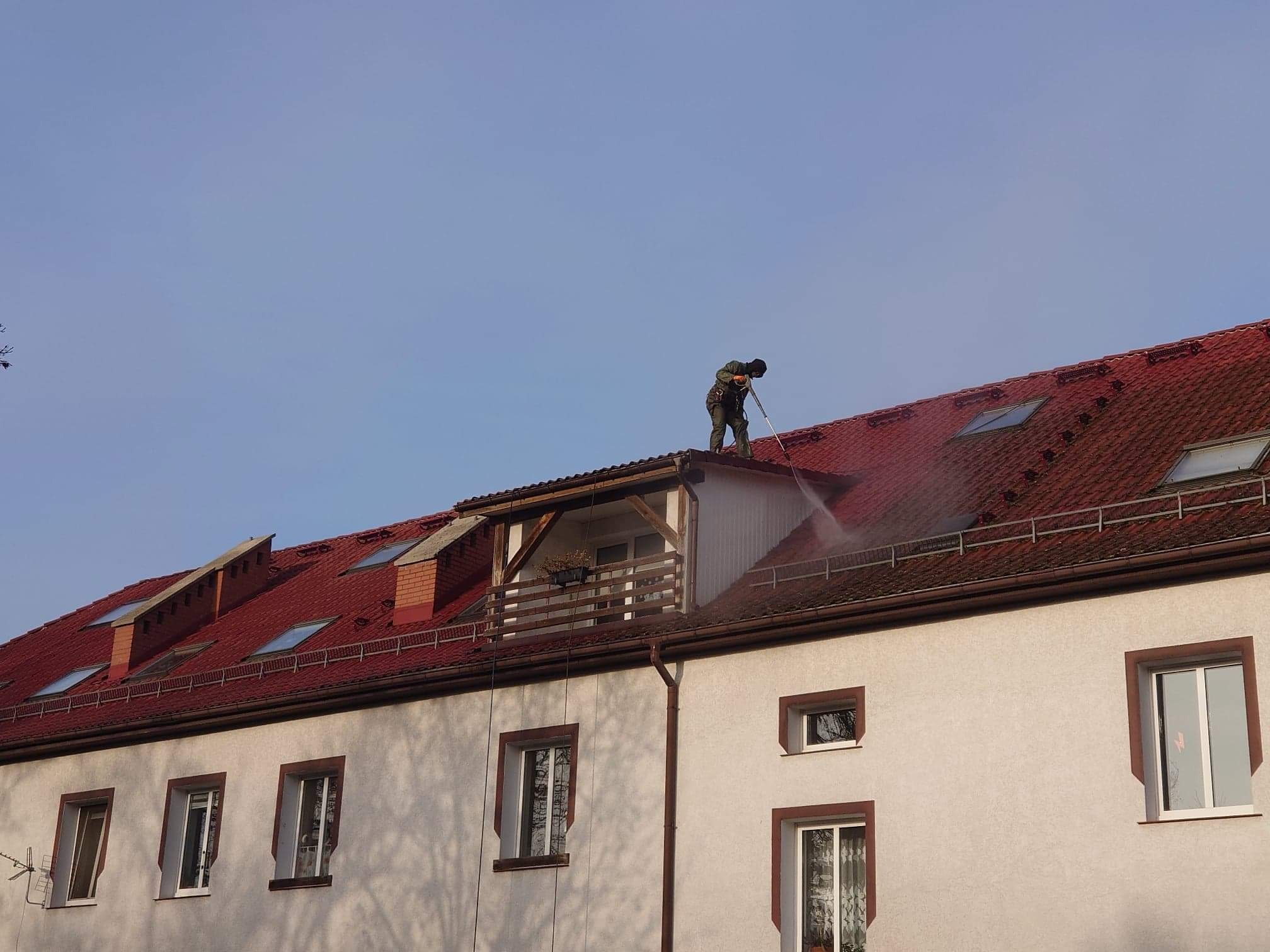 Pracownik w kombinezonie ochronnym czyści czerwoną dachówkę budynku wielorodzinnego za pomocą myjki ciśnieniowej, widoczne okna dachowe i balkon.