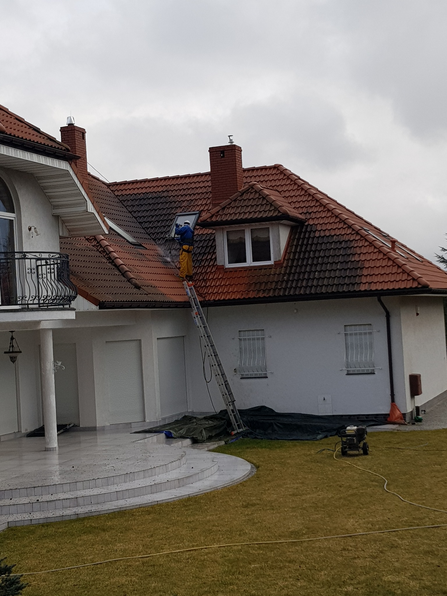 Pracownik w niebieskiej kurtce i żółtych spodniach na drabinie, czyści dach z czerwonej dachówki za pomocą myjki ciśnieniowej; widoczna różnica w czystości dachu przed i po czyszczeniu; duży, biały...