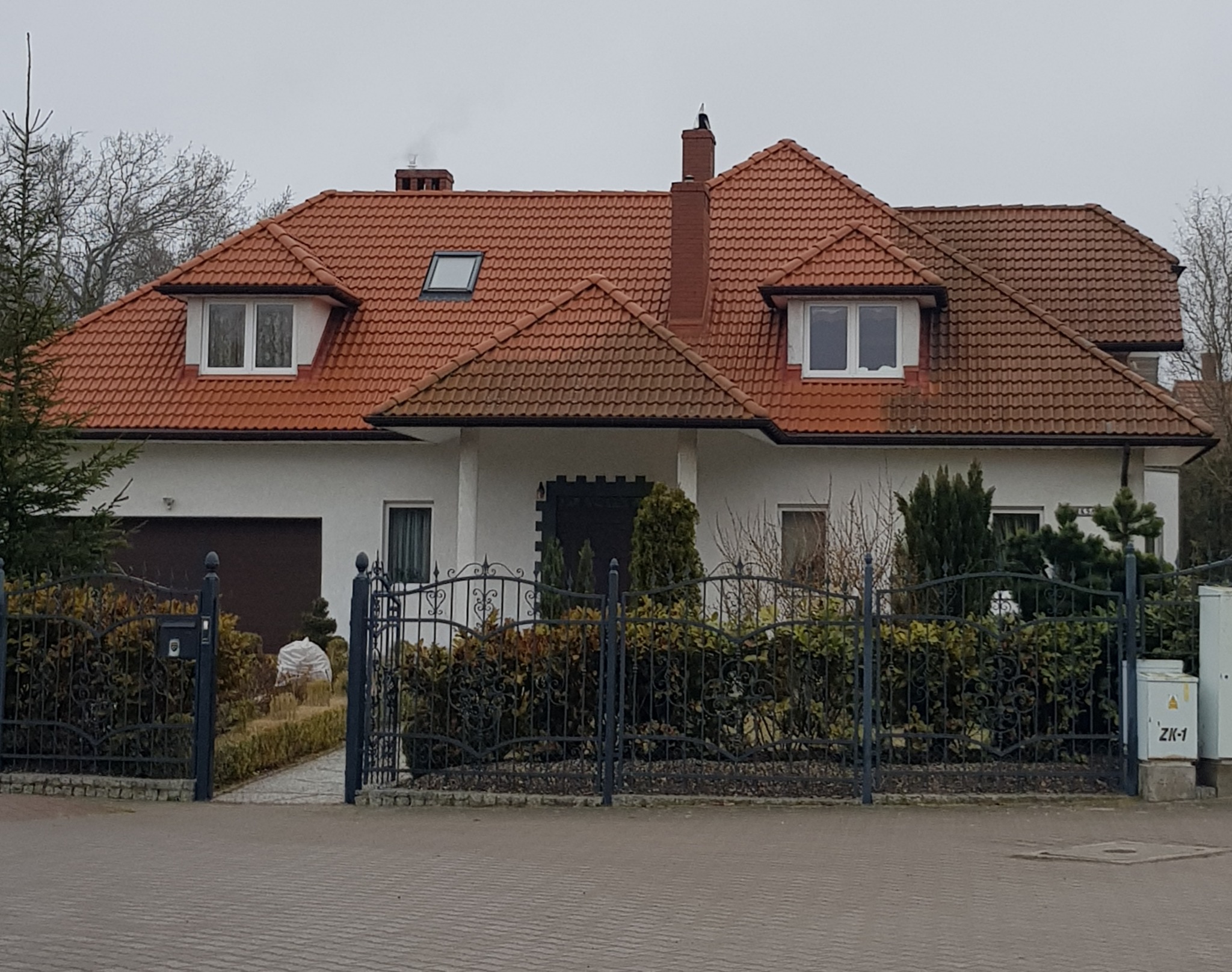 Dom jednorodzinny z czerwoną dachówką, widoczny zza ozdobnego, czarnego ogrodzenia. Na dachu komin z ptakiem, okna dachowe i lukarny. Elewacja biała.