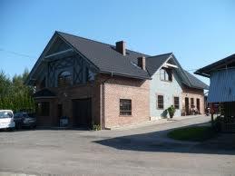 Dwukondygnacyjny dom z ceglaną elewacją i ciemnym dachem. Część budynku pomalowana na szaro. Widoczny podjazd i fragment otoczenia z drzewami.