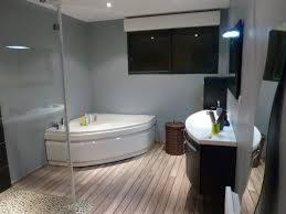 Nowoczesna łazienka z narożną wanną, drewnopodobną podłogą i szklaną kabiną prysznicową. Minimalistyczny wystrój, ciemne okno i szafka pod umywalką.