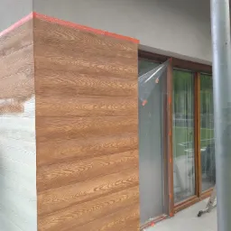 Fragment elewacji budynku w trakcie prac wykończeniowych: część ściany z imitacją drewna w kolorze brązowym, druga część pomalowana na biało, okno zabezpieczone folią malarską, widoczne ślady farby...