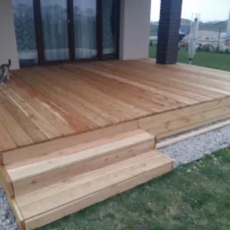 Nowoczesny taras z drewna z trzema schodami, otoczony trawnikiem i szarym żwirem; kot w rogu tarasu.