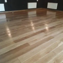 Błyszcząca, jasna podłoga parkietowa z wyraźnym usłojeniem, ułożona w jodełkę, odbijająca światło w przestronnym wnętrzu z ciemną ścianą i białymi grzejnikami.