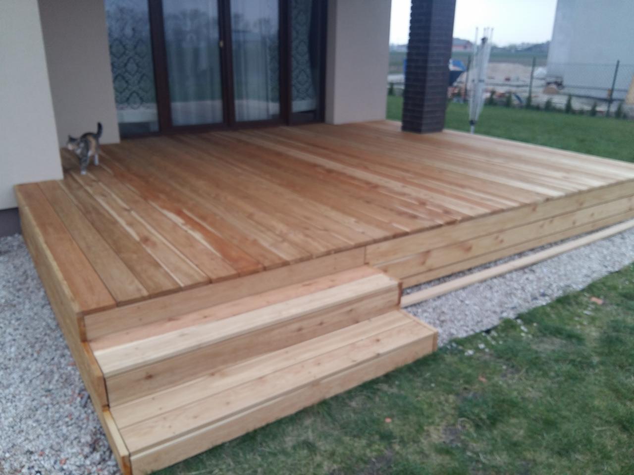 Nowoczesny taras z drewna z trzema schodami, otoczony trawnikiem i szarym żwirem; kot w rogu tarasu.