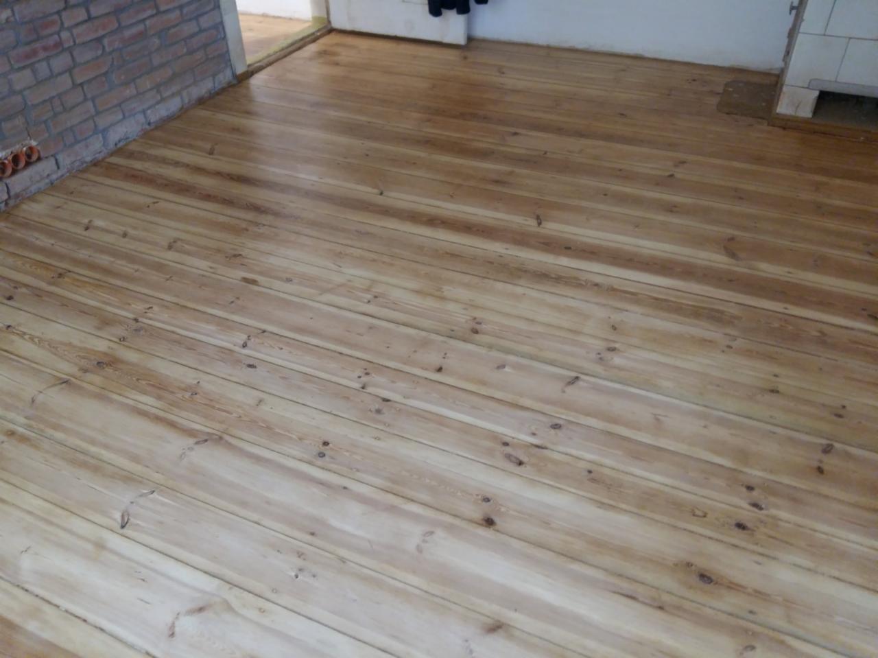 Świeżo wycyklinowana, jasna podłoga drewniana z widocznymi słojami i fragmentem ściany z cegły w tle.