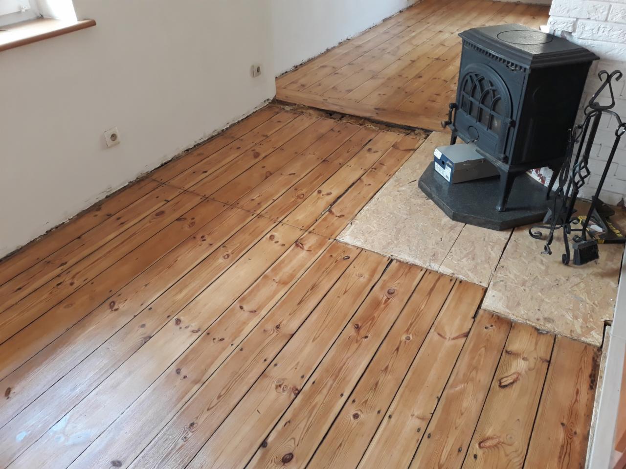 Wnętrze z drewnianą podłogą po renowacji, widoczne ślady po cyklinowaniu i zabezpieczaniu lakierem, piec wolnostojący na tle białej ściany.