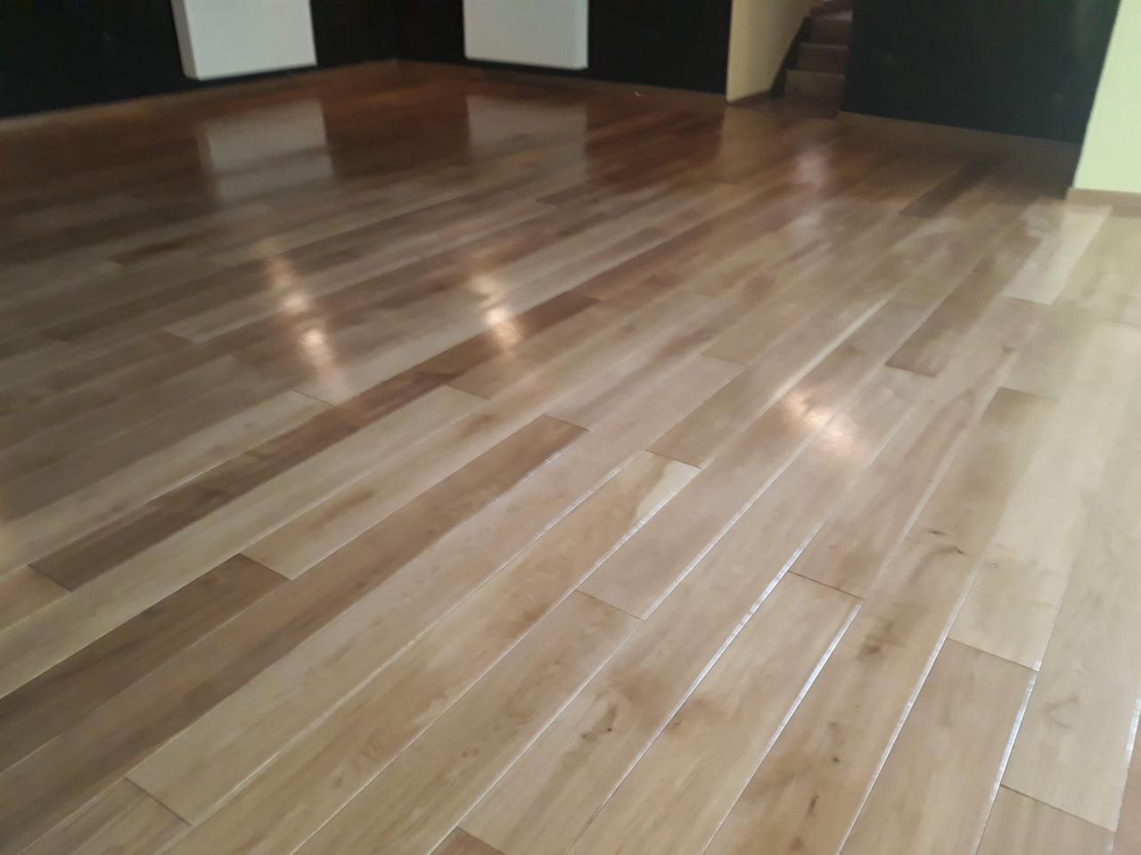 Błyszcząca, jasna podłoga z drewnianych paneli ułożonych wzdłużnie, z widocznymi refleksami światła i fragmentem schodów w tle.