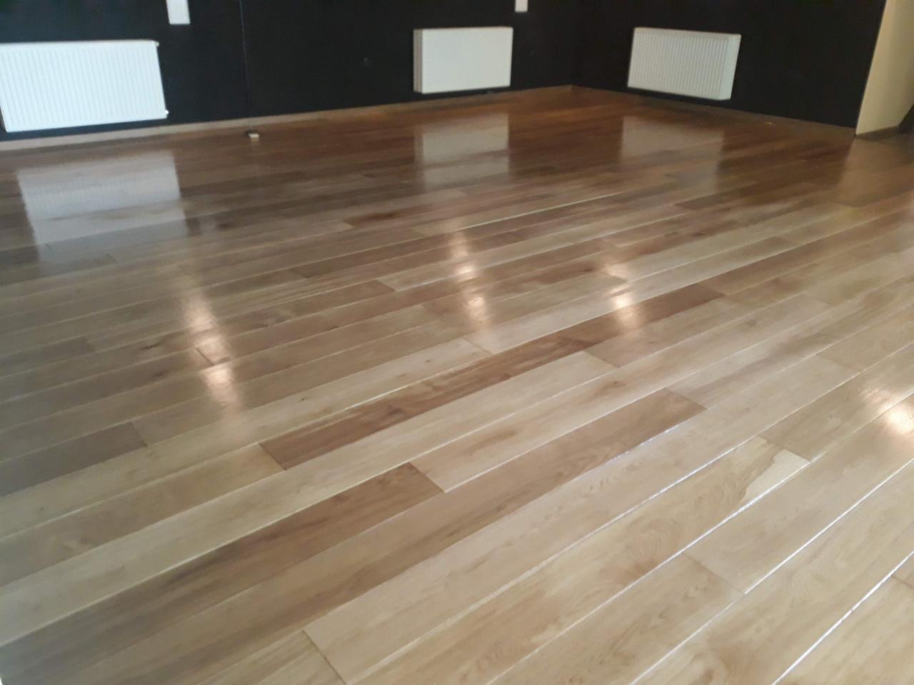 Błyszcząca, jasna podłoga parkietowa z wyraźnym usłojeniem, ułożona w jodełkę, odbijająca światło w przestronnym wnętrzu z ciemną ścianą i białymi grzejnikami.