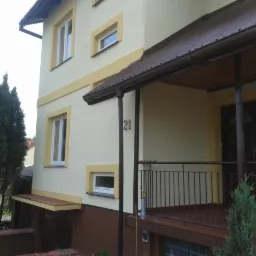 Odnowiona elewacja domu z numerem 21, kremowa z ż&oacute;łtymi akcentami, brązowy cok&oacute;ł, widoczne okna i zadaszony ganek z metalową balustradą.