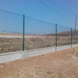 Długi płot z siatki, zielone słupki, betonowa podmurówka, za płotem pole i drzewa, błękitne niebo. Widok z dołu, widać kable elektryczne.