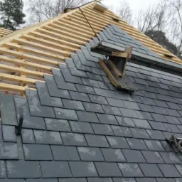 Montaż łupka na dachu: widok na drewnianą więźbę, częściowo pokrytą szarym łupkiem, z metalową platformą roboczą i drabiną, w tle drzewa bez liści i fragment komina.