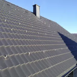 Nowy, grafitowy dach z dachówki ceramicznej z zamontowanymi klamrami mocującymi, widoczny komin i fragment ściany budynku w surowym stanie.