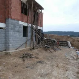 Budowa domu w stanie surowym otwartym: ściany z czerwonej cegły i szarego bloczka, drewniane stemple podtrzymujące strop, widoczna betoniarka w tle na nierównym terenie.