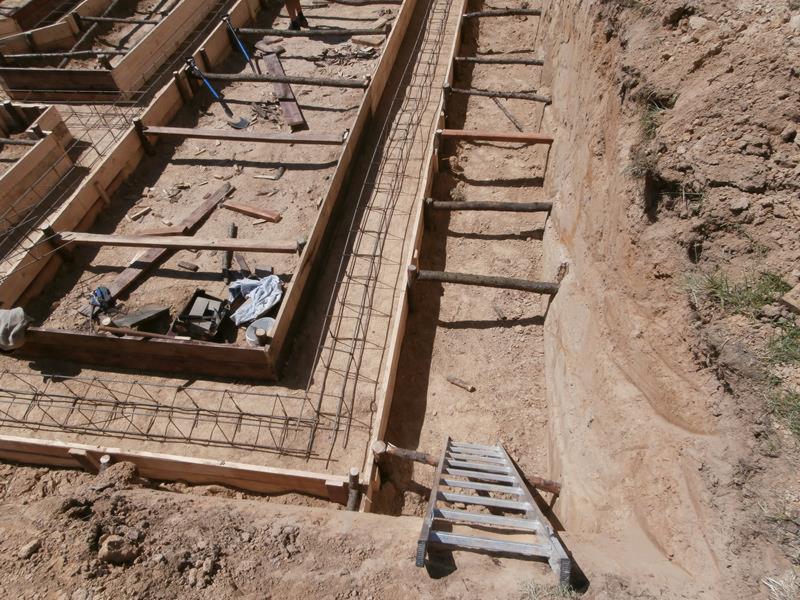 Budowa fundamentów: wykop z drewnianą drabinką, szalunki zbrojeniowe z siatki stalowej, widoczne narzędzia i fragment ubrania roboczego.