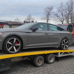 F.H.U. KAMIR - Szary samochód Audi na żółtej lawecie, z widocznymi czerwonymi zaciskami hamulcowymi i czarnymi felgami, transportowany na tle budynku i drzew bez liści.