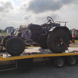 F.H.U. KAMIR - Żółta laweta z czarnym traktorem na platformie, zabezpieczonym pasami. W tle pole, zabudowania gospodarcze i pochmurne niebo. Widok z boku, lekko z dołu.
