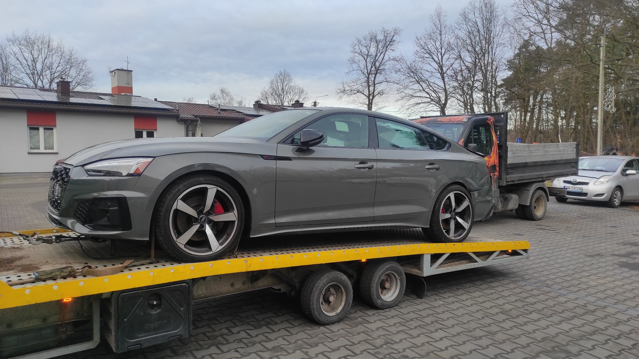 Szary samochód Audi na żółtej lawecie, z widocznymi czerwonymi zaciskami hamulcowymi i czarnymi felgami, transportowany na tle budynku i drzew bez liści.