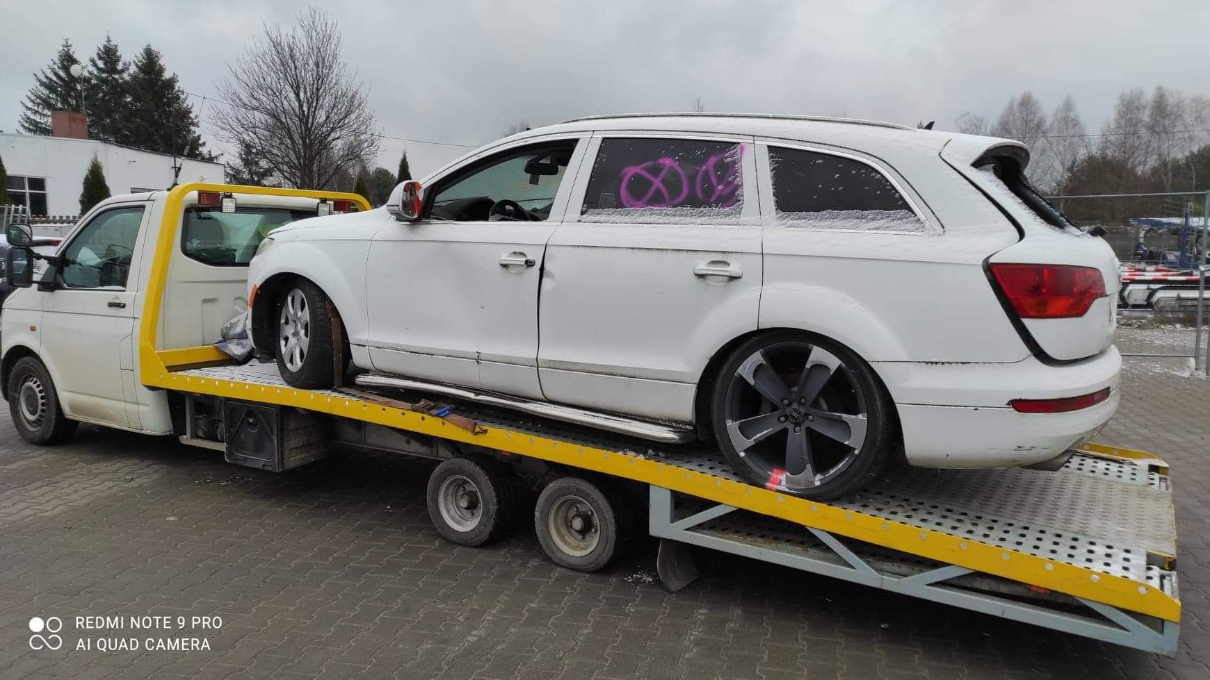 Białe Audi Q7 z pomalowanymi na różowo szybami leży na żółtej platformie lawety, widok z boku na tle szarej kostki brukowej i budynków.