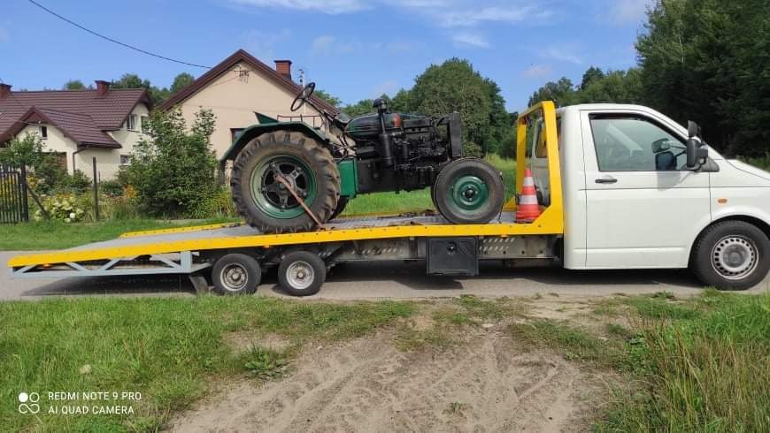 Zielony, zabytkowy traktor zabezpieczony na żółtej platformie lawety, transportowany białą furgonetką na tle wiejskiego krajobrazu.