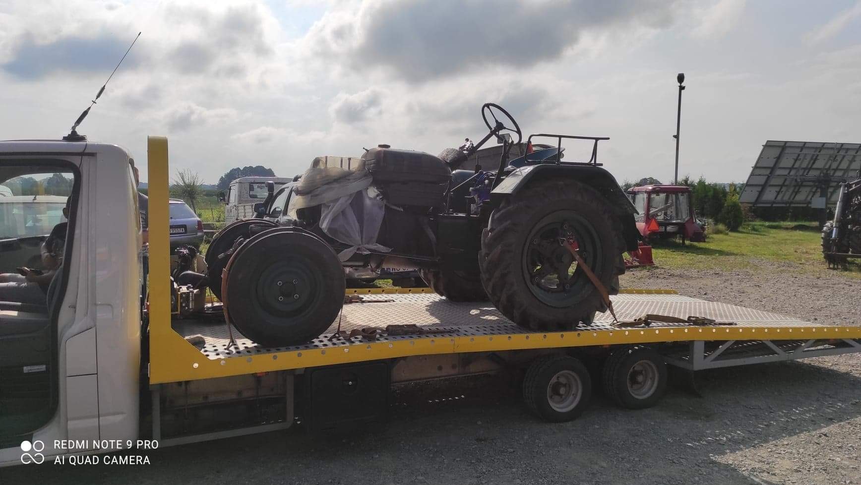 Żółta laweta z czarnym traktorem na platformie, zabezpieczonym pasami. W tle pole, zabudowania gospodarcze i pochmurne niebo. Widok z boku, lekko z dołu.