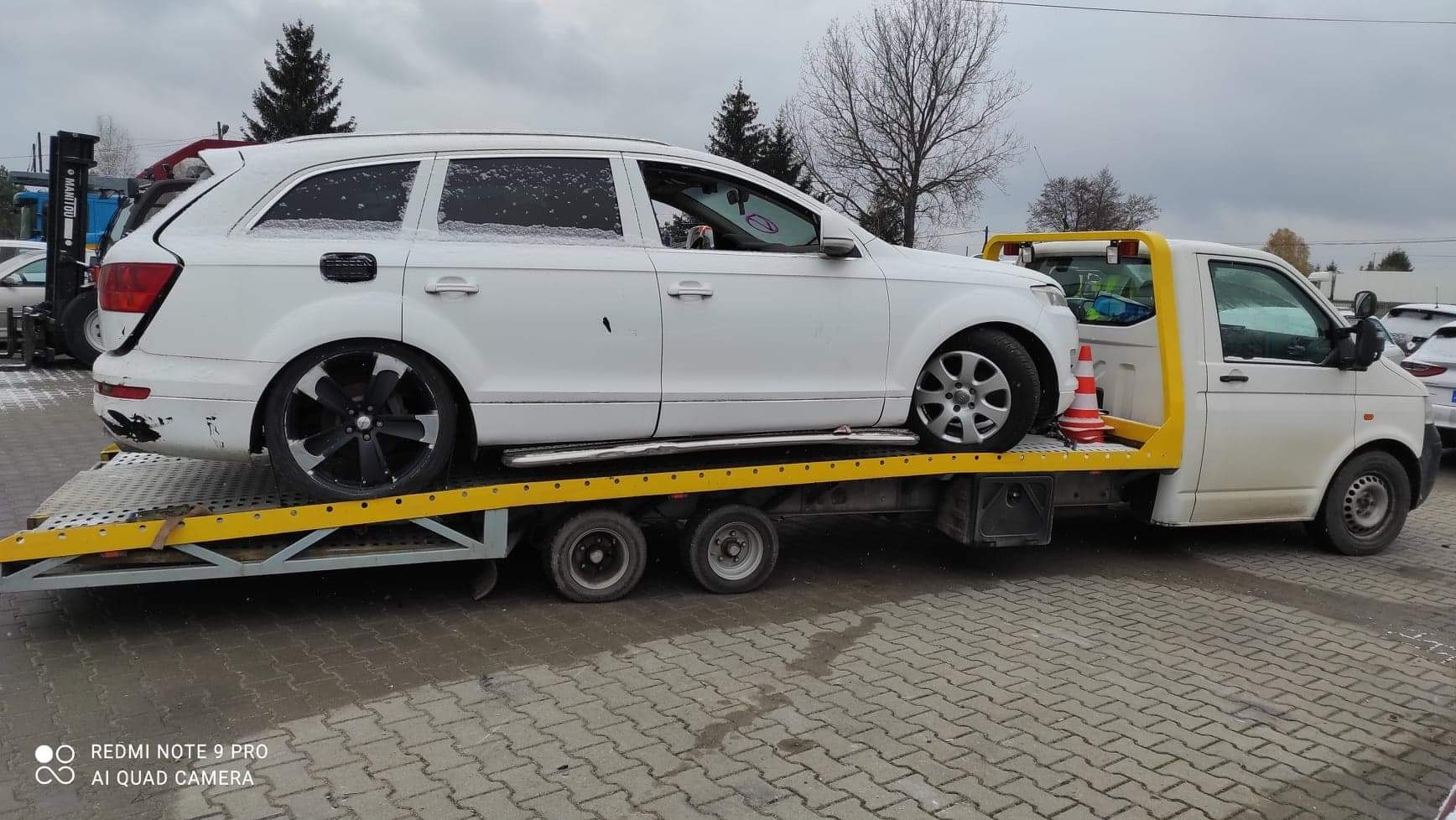 Biały samochód osobowy Audi Q7 na żółtej lawecie, widok z boku na szarym brukowanym placu. Na aucie widoczny śnieg.