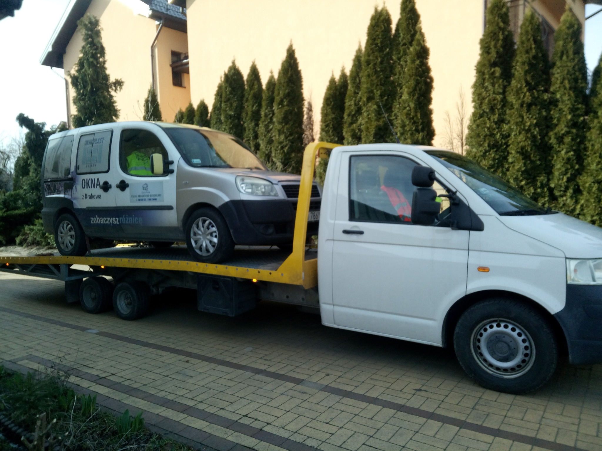 Laweta z załadowanym busem firmy OKNA z Krakowa na tle zielonych tuj i budynku. Żółty najazd, biała kabina lawety. Transport na brukowanej drodze.