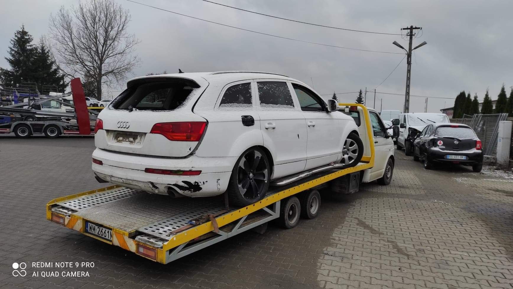 Białe Audi Q7 z uszkodzeniami na żółtej lawecie, widok z tyłu, lekko ośnieżone. Tablica rejestracyjna lawety widoczna. W tle inne pojazdy i słup energetyczny.