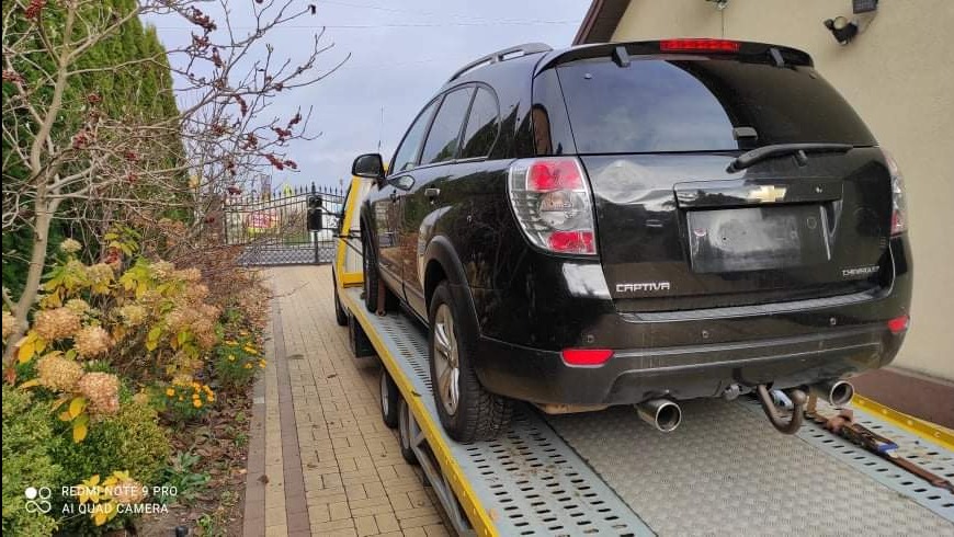 Czarny Chevrolet Captiva na żółtej lawecie, widok z tyłu, na tle domu i ogrodu. Hak holowniczy i liny zabezpieczające widoczne na platformie.