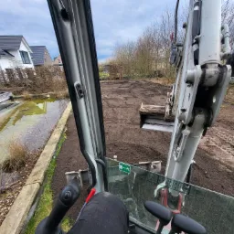 Widok z kabiny minikoparki na wyrównywany teren pod przyszły ogród, z widocznym stawem w tle i domem z panelami słonecznymi po lewej stronie.