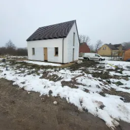 Surowy, biały dom z brązowym dachem w zimowym krajobrazie. Widoczny samochód dostawczy i ślady prac budowlanych. Minimalistyczna architektura w trakcie wykończenia.
