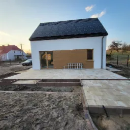 Nowoczesny dom z panelami słonecznymi i częściowo ułożonym tarasem z kostki. Widoczne przygotowane podłoże pod dalszą część tarasu. Drabina oparta o ścianę.
