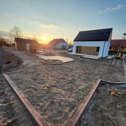 Garden Mission Possible Twój ogród to nasza misja - Etap prac w ogrodzie: obrzeża z kostki, przygotowane podłoże pod trawnik, widoczny dom z panelami słonecznymi w tle, zachód słońca.