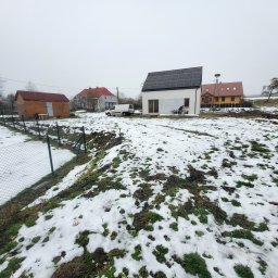 Garden Mission Possible Twój ogród to nasza misja - Nowoczesny dom z panelami słonecznymi na dachu, widok z zasypanego śniegiem ogrodu. W tle inne zabudowania i częściowo widoczny samochód dostawczy.