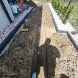 Garden Mission Possible Twój ogród to nasza misja - Głęboki wykop pod instalację drenażową lub nawadniającą, z żółtą rurą, wzdłuż ściany budynku i rabaty z białym grysem i tujami.