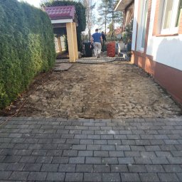Garden Mission Possible Twój ogród to nasza misja - Częściowo ułożona ciemnoszara kostka brukowa tworząca ścieżkę, przechodząca w przygotowany grunt obok zielonego żywopłotu i białej ściany domu. Widoczny pomarańczowy kabel.