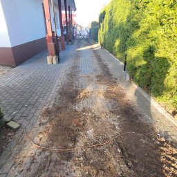 Garden Mission Possible Twój ogród to nasza misja - Zabrudzona kostka brukowa na podjeździe, pokryta błotem i drobnymi kamieniami, prowadząca wzdłuż ściany domu i gęstego, zielonego żywopłotu. Na ziemi leży pomarańczowy przewód.