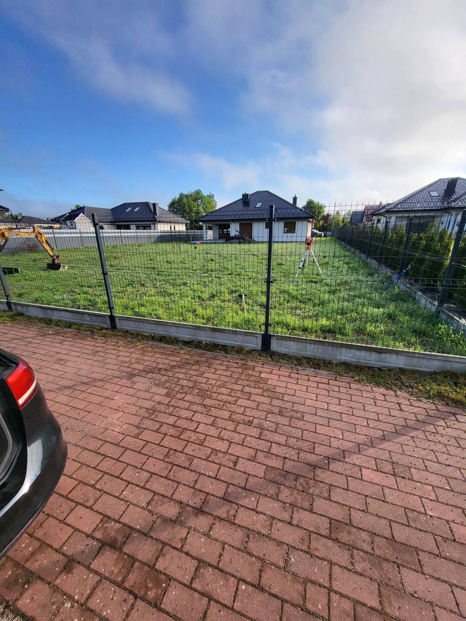 Nowoczesne ogrodzenie panelowe z podmurówką betonową oddzielające posesję z trawnikiem, widoczny niwelator i minikoparka w tle, kostka brukowa na pierwszym planie.